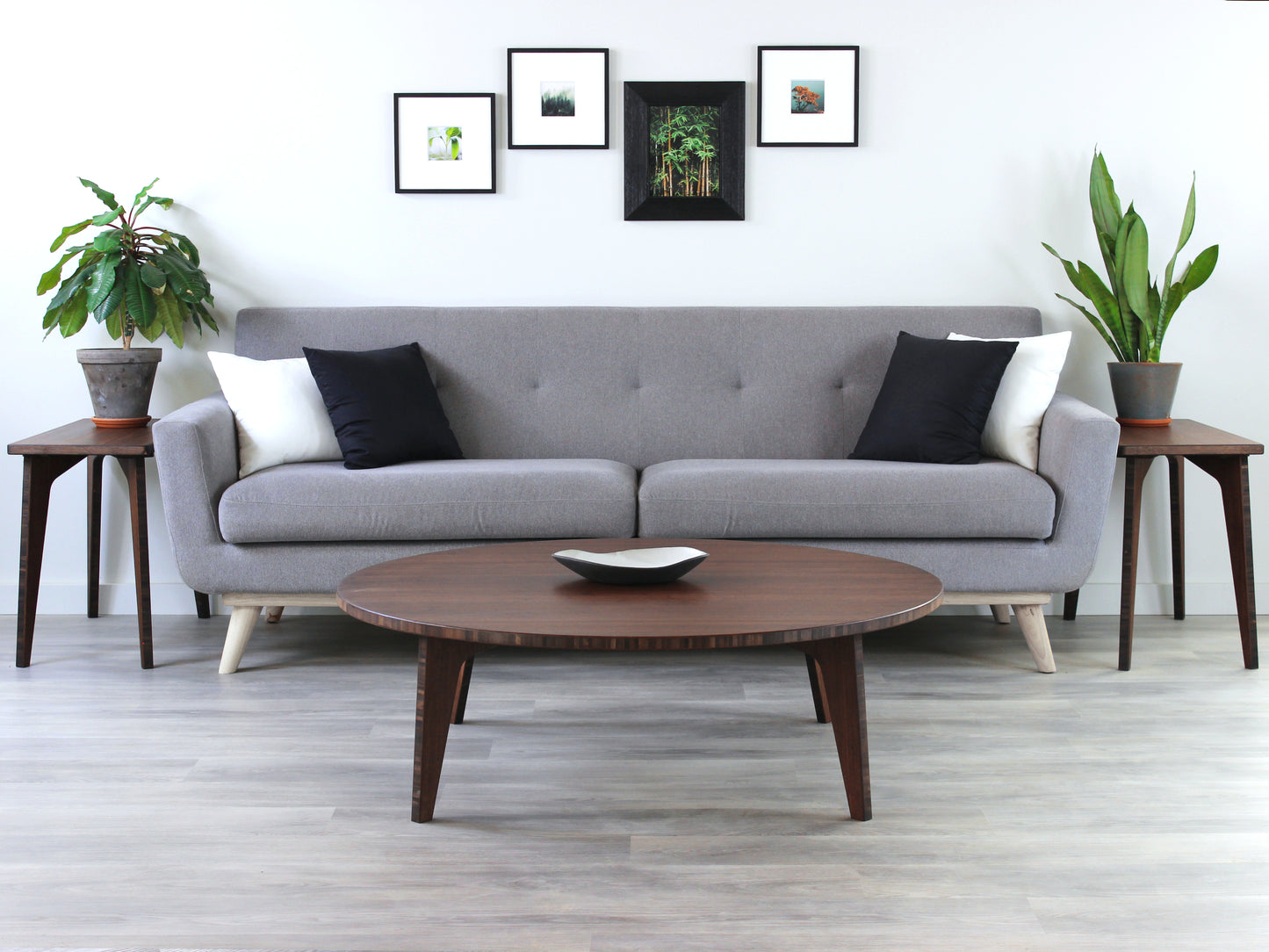 Large circle floor table in bamboo with a walnut color finish, featuring a Japandi style design, set on a grey floor with a white background. Set in a living room area, a grey couch and side tables with houseplants. A small black pottery vessel is placed on the table top.