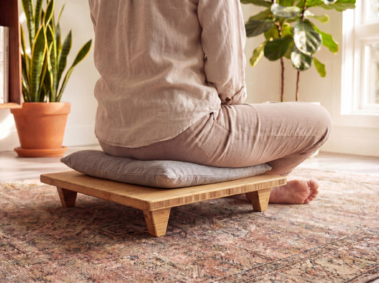 Person sitting cross-legged on a bamboo platform with cushion in a room with plants and a rug.

Meditation Chair 
Japanese Tea Table 
Kotatsu
low chair
posture support 
ergonomic 
minimilist 