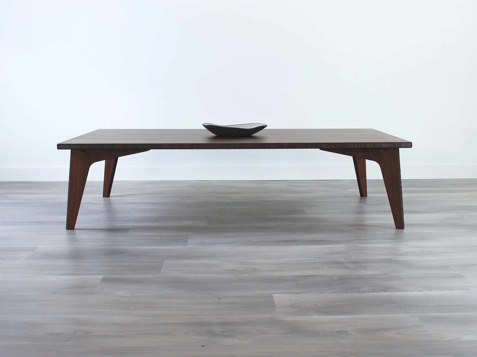 A large rectangular low table made of bamboo with a walnut finish, placed on a grey floor, designed for multi-functional use in living spaces. A small black pottery vessel set on the table top. 

japanese furniture
japanese low table
low coffee table
low dining table
low table
mid century
minimalist
minimalist table
modern