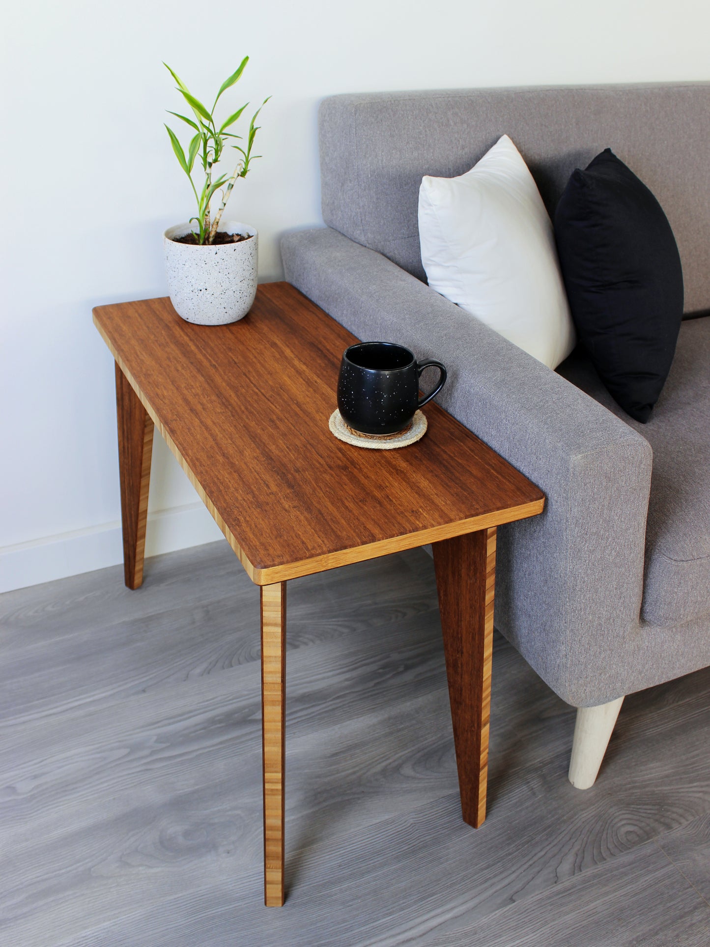 A rectangular side table with a chestnut finish bamboo top and legs, placed on a grey floor, with a white background. Black cup and small house plant on top of the table. Side table next to a grey couch with black and white pillows.
mid century
minimalist
chabudai
coffee table
side table