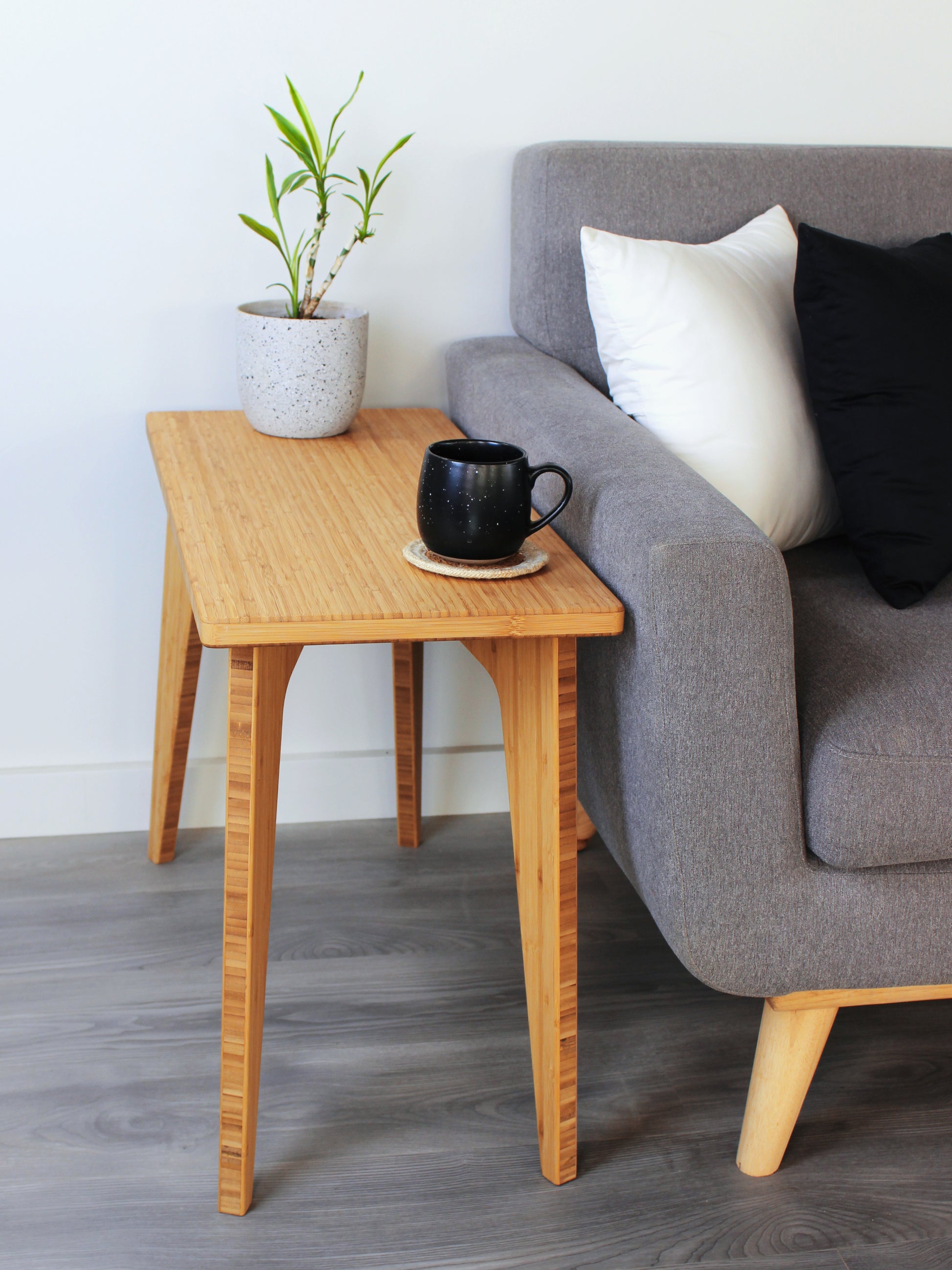A rectangular side table with a natural finish bamboo top and legs, placed on a grey floor, with a white background. Black cup  and small house plant on top of the table. Side table next to a grey couch with black and white pillows.
mid century
minimalist
chabudai
coffee table
side table