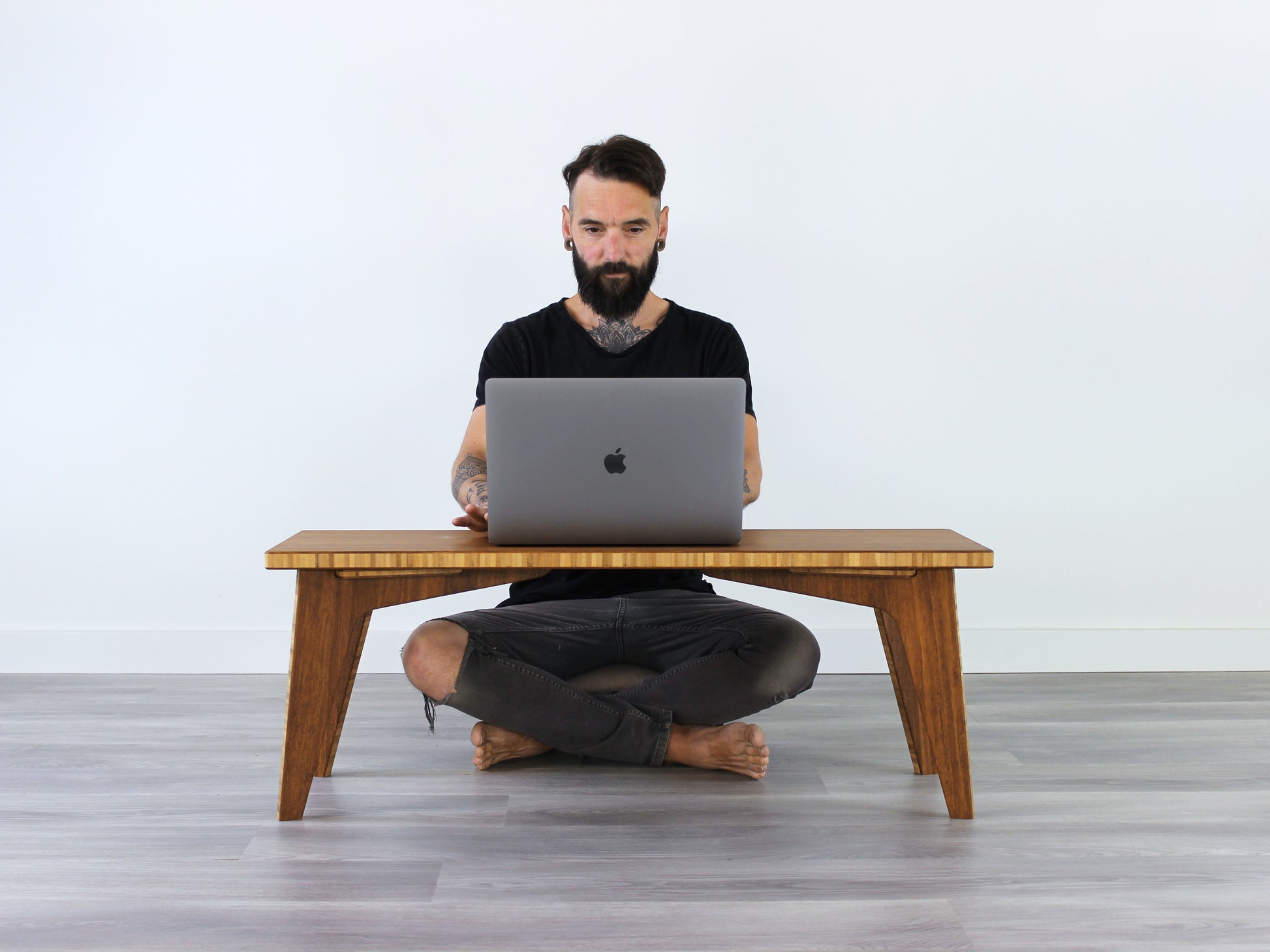 A small rectangular floor desk made of bamboo with a chestnut finish, featuring a person sitting cross legged, working on a laptop.Situated on a grey floor with a white wall in the background.
japanese furniture
japanese low table
mid century
minimalist
minimalist table
low desk 
computer desk 
laptop table 