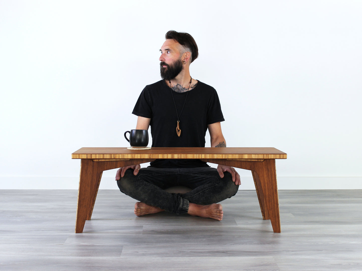 A small rectangular floor desk made of bamboo with a chestnut finish, featuring a person sitting looking to the left. A black mug is sitting on the table top, all is placed on a grey floor with a white wall in the background. 

mid century, minimalist, minimalist table, modern, square coffee table, table, tatami, Floor chair with back, posture support, low chair, chestnut bamboo