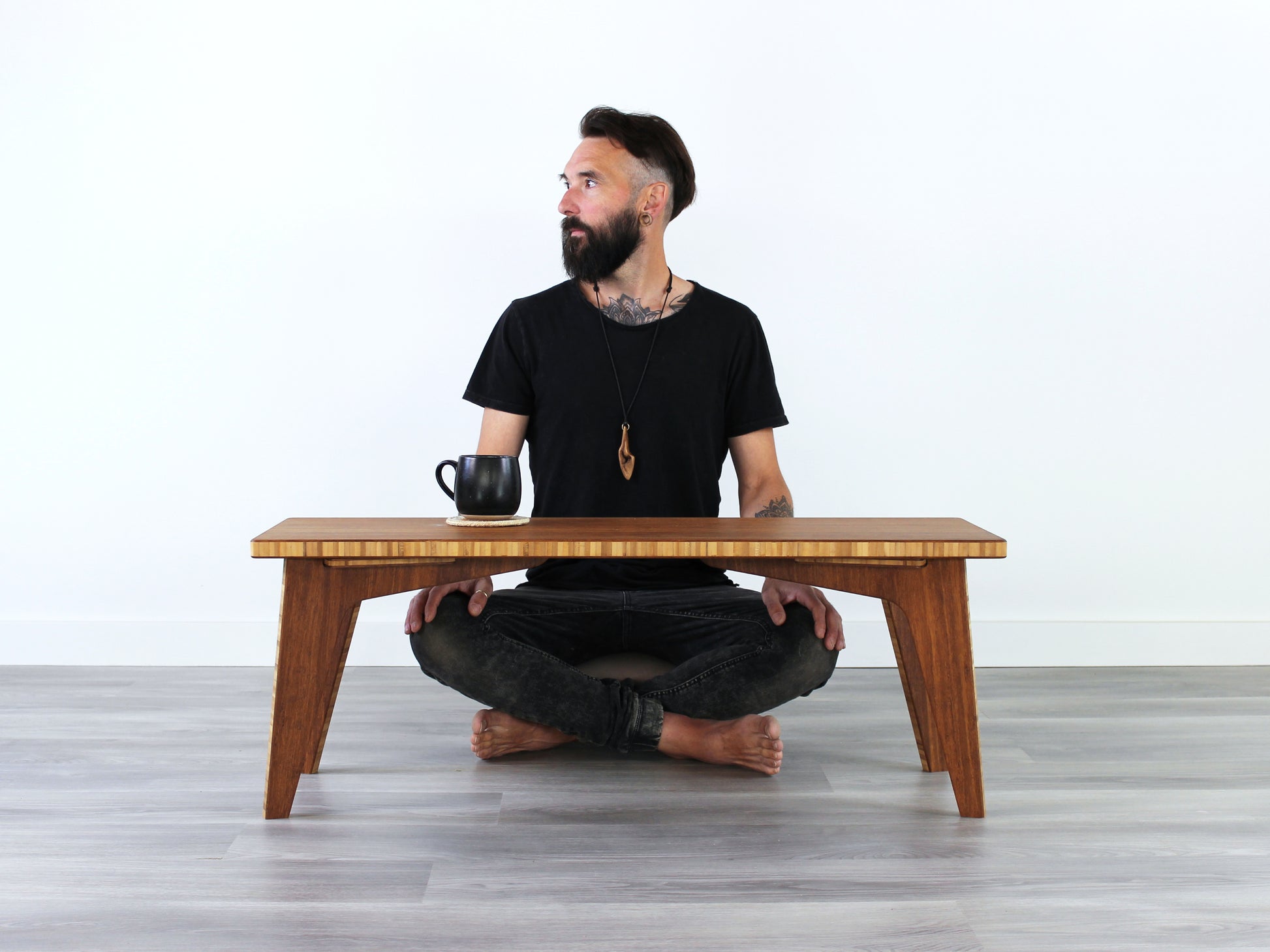 A small rectangular floor desk made of bamboo with a chestnut finish, featuring a person sitting looking to the left. A black mug is sitting on the table top, all is placed on a grey floor with a white wall in the background. 

mid century, minimalist, minimalist table, modern, square coffee table, table, tatami, Floor chair with back, posture support, low chair, chestnut bamboo