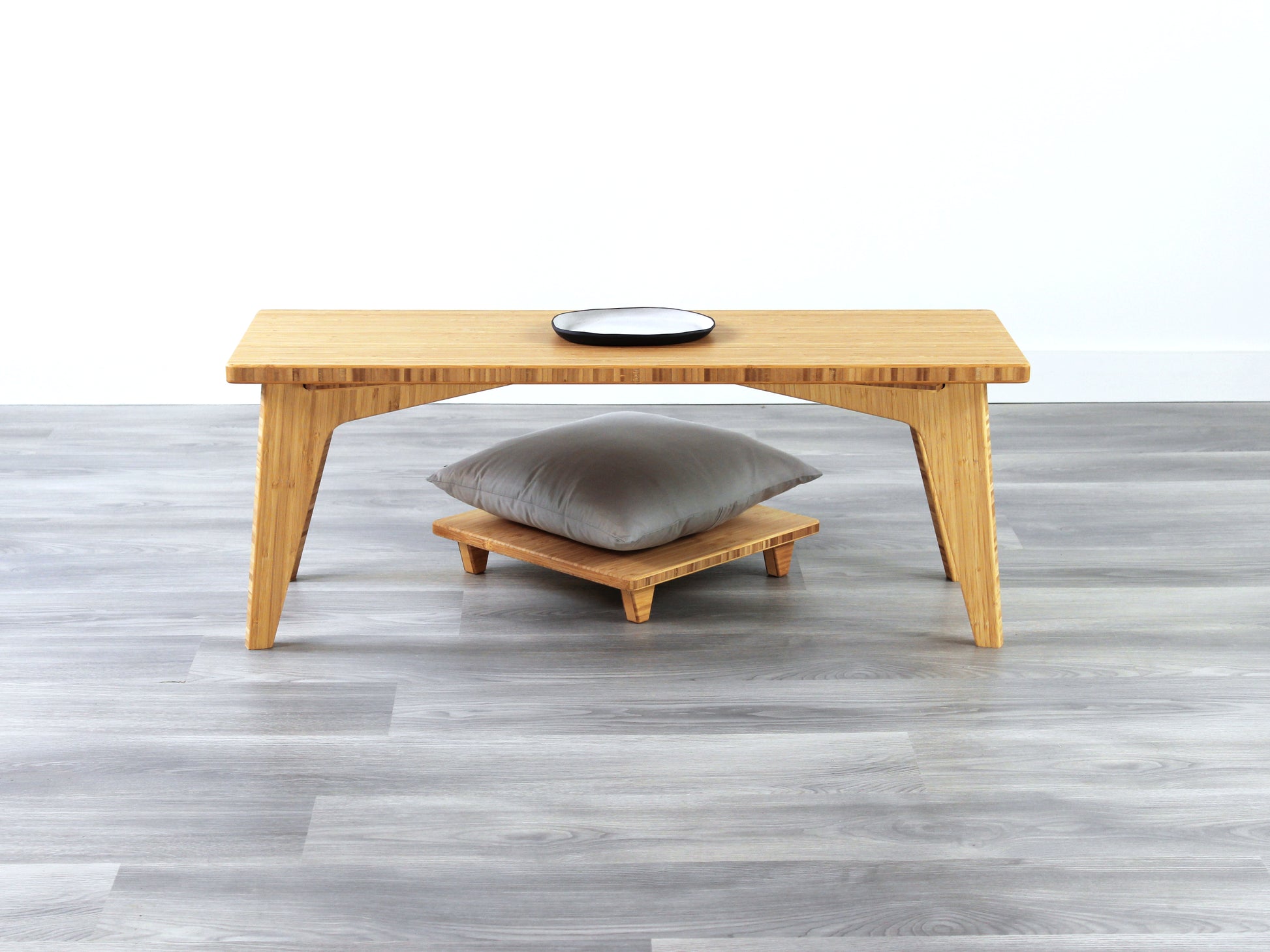 A small rectangular floor desk made of bamboo with a natural finish, featuring a single black piece of pottery on its top surface, placed on a grey floor with a white wall in the background. Floor Chair Posture placed beneath.

mid century, minimalist, minimalist table, modern, square coffee table, table, tatami, Floor chair, posture support, low chair, chestnut bamboo