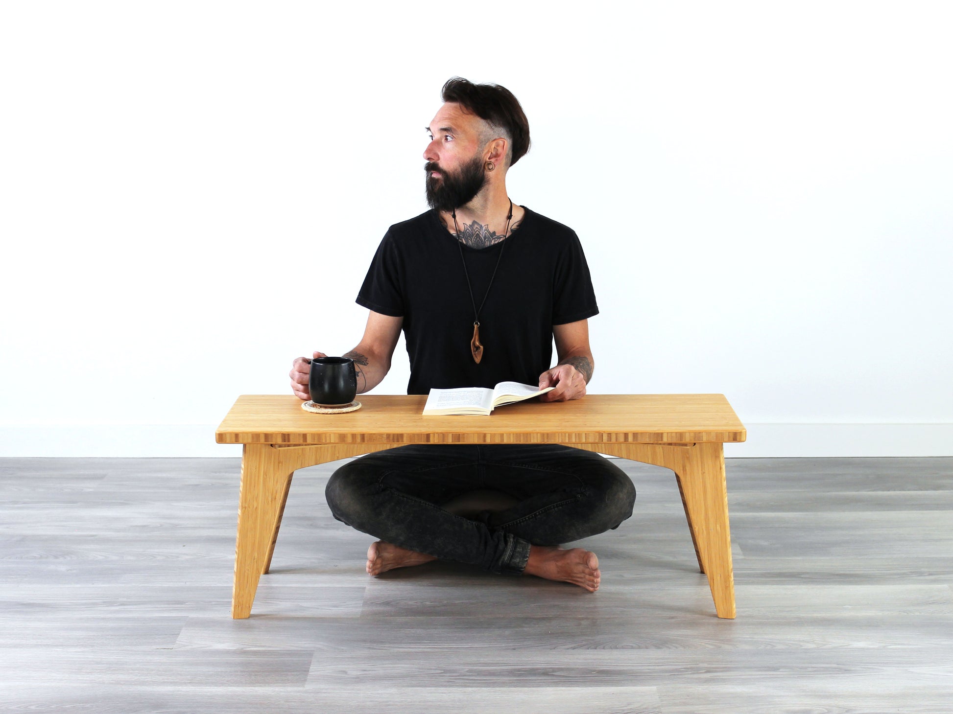 A small rectangular floor desk made of bamboo with a natural finish, featuring a person sitting looking to the left. A black mug is sitting on the table top, all is placed on a grey floor with a white wall in the background. 

mid century, minimalist, minimalist table, modern, square coffee table, table, tatami, Floor chair with back, posture support, low chair, chestnut bamboo
