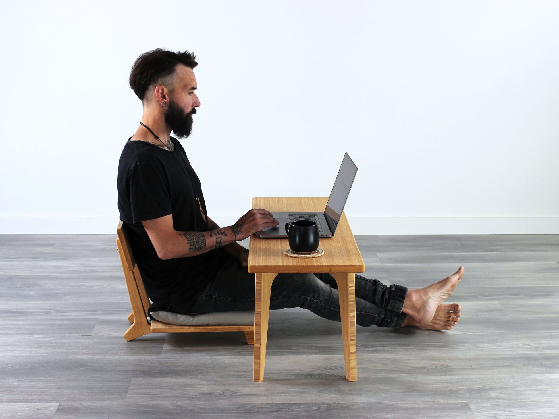 A small rectangular floor desk made of bamboo with a natural finish, featuring a person sitting in a Floor Chair Comfort working on a laptop. Placed on a grey floor with a white wall in the background. 

mid century, minimalist, minimalist table, modern, square coffee table, table, tatami, Floor chair with back, posture support, low chair, chestnut bamboo