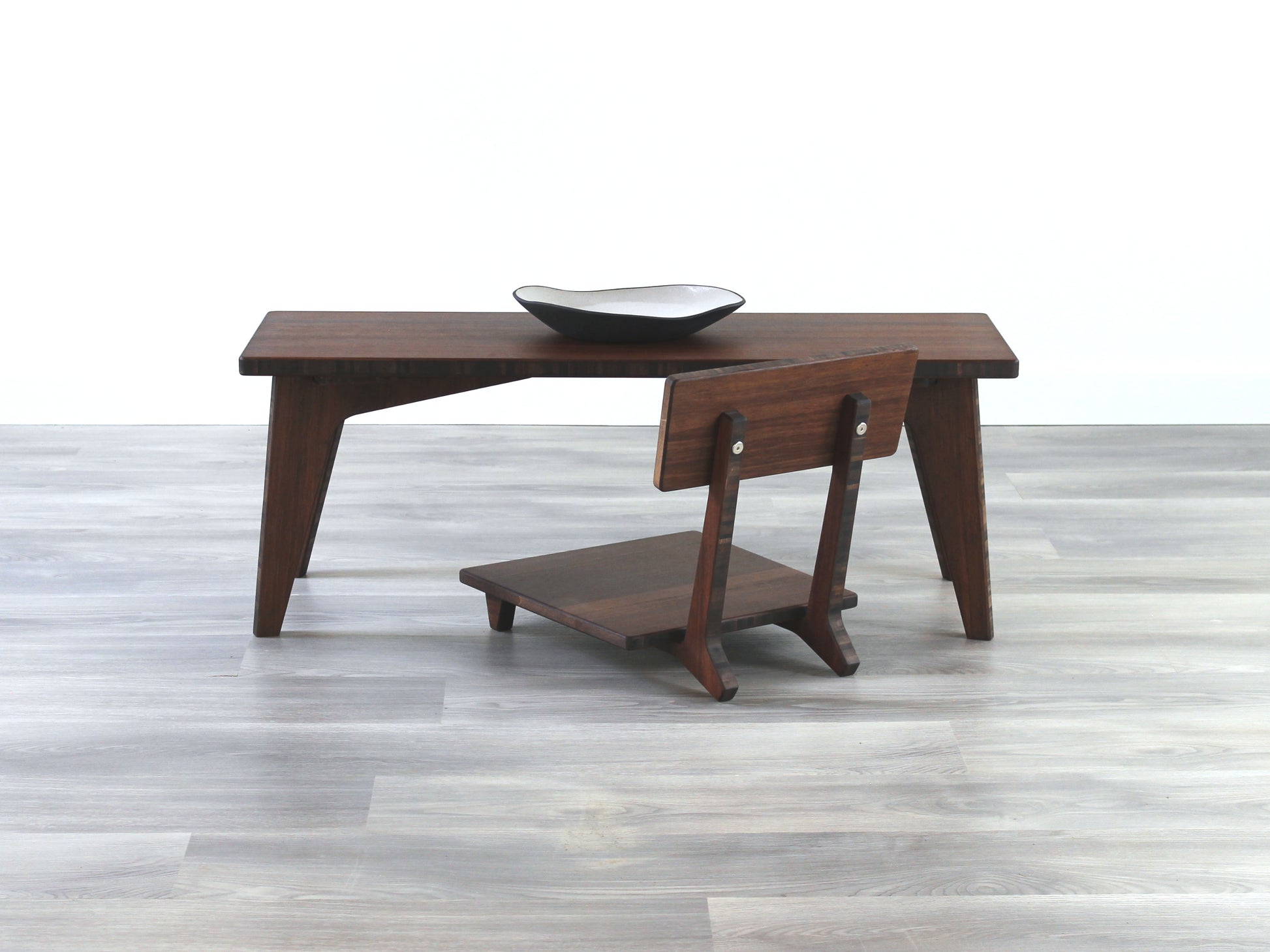 A small rectangular floor desk made of bamboo with a walnut finish, featuring a single black piece of pottery on its top surface, placed on a grey floor with a white wall in the background. Floor Chair Comfort placed beneath.


walnut bamboo coffee table, low chair chabudai, tatami, japanese, mid-century, organic bowl on table