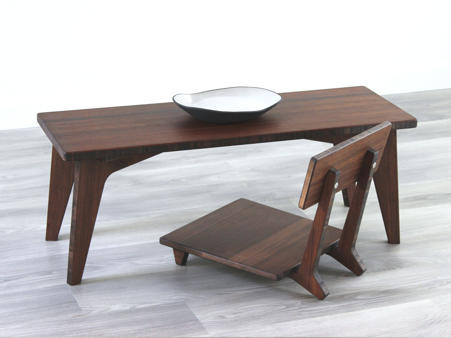 A small rectangular floor desk made of bamboo with a walnut finish, featuring a single black piece of pottery on its top surface, placed on a grey floor with a white wall in the background. Floor Chair Comfort placed beneath.


walnut bamboo coffee table, low chair with back, chabudai, tatami, japanese, mid-century, organic bowl on table