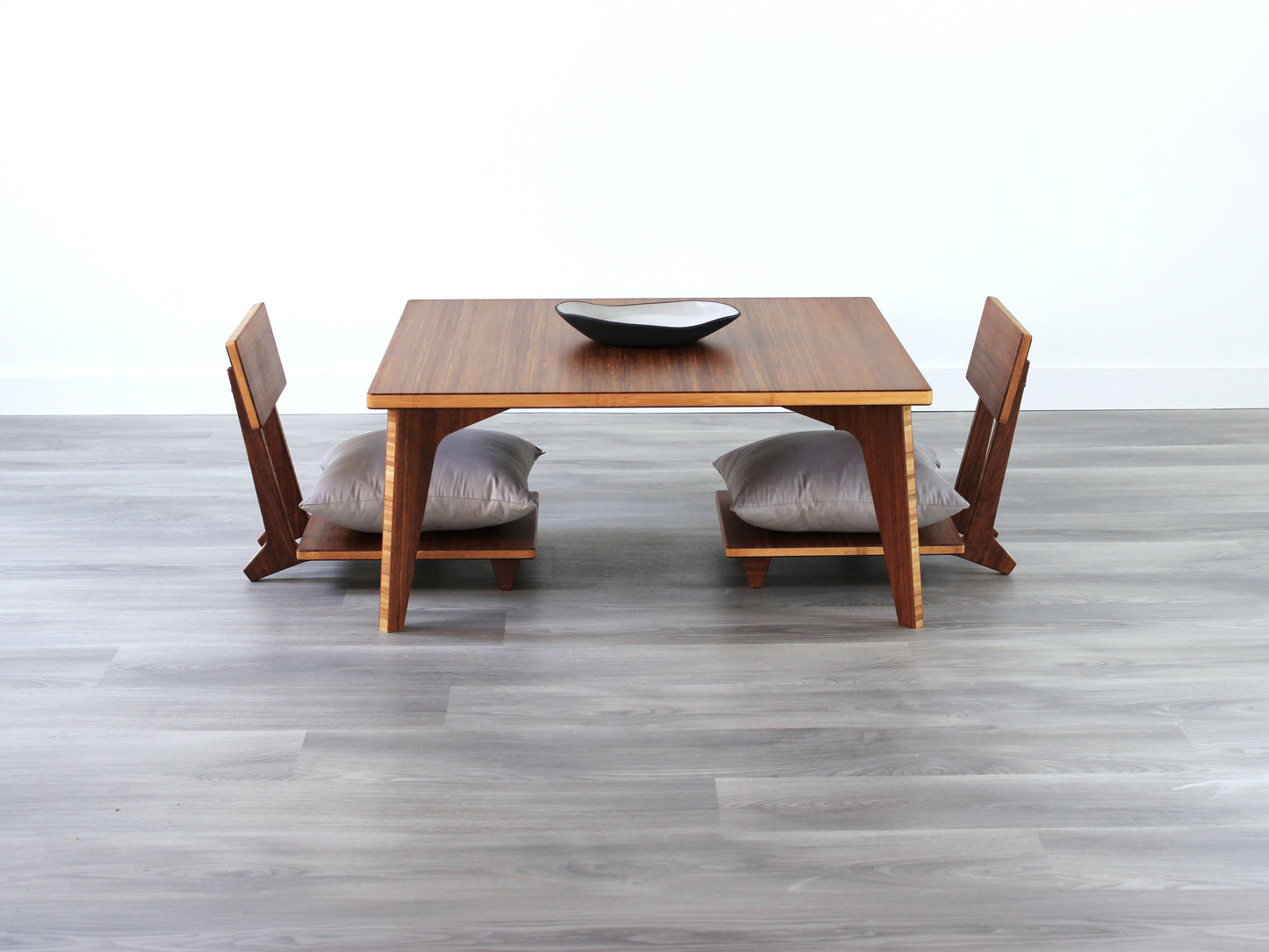 Small Square Table Set in chestnut finish. Two Floor Chair Comfort with grey pillows are placed below. A small black pottery vessel is placed on top of the table. 

sustainable bamboo, renewable bamboo, solid bamboo, eco-friendly furniture, zero-waste, carbon neutral, natural materials, natural-based finish, repairable furniture