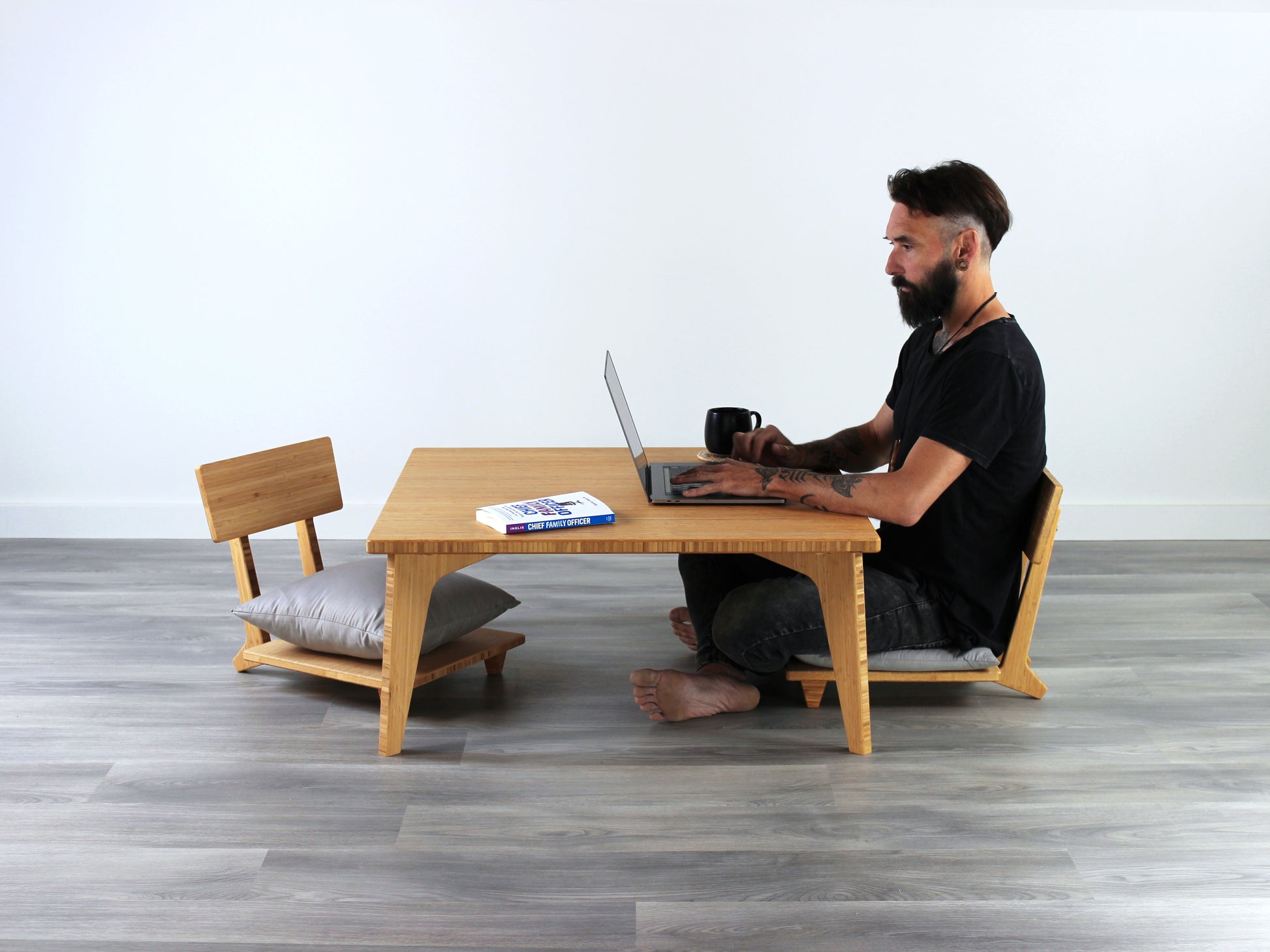 Small Square Table Set in natural finish. Two Floor Chair Comfort with grey pillows are placed below. A person is sitting in one and working on a laptop. A black coffee mug is placed on top of the table. 

sustainable bamboo, renewable bamboo, solid bamboo, eco-friendly furniture, zero-waste, carbon neutral, natural materials, natural-based finish, repairable furniture