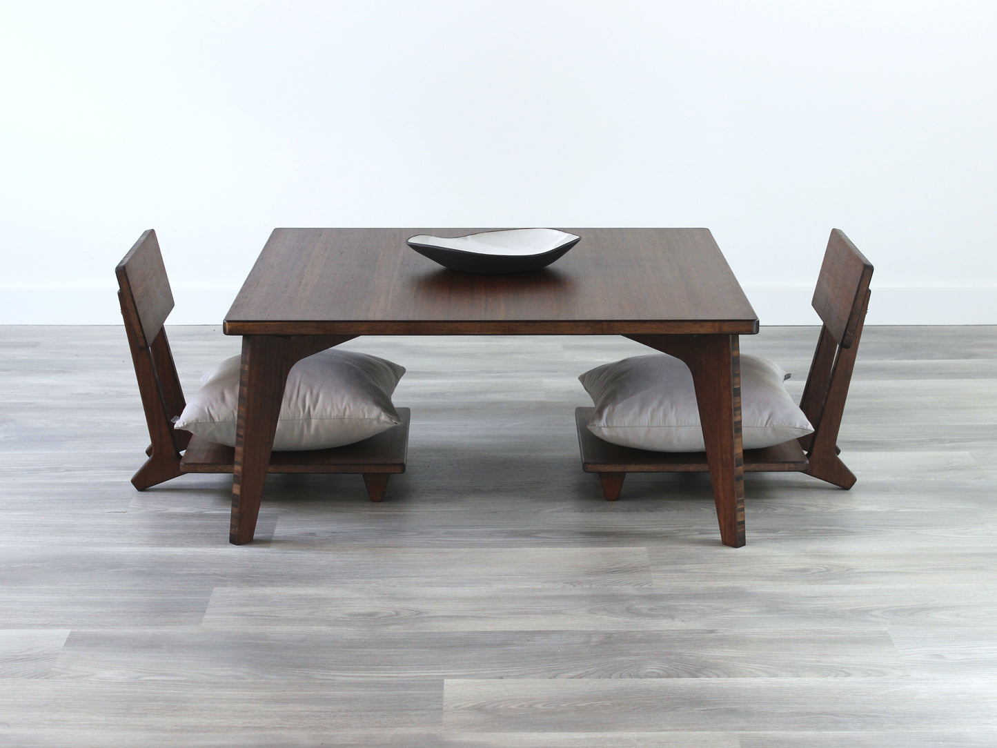 Small Square Table Set in walnut finish. Two Floor Chair Comfort with grey pillows are placed below. A small black pottery vessel is placed on top of the table. 

sustainable bamboo, renewable bamboo, solid bamboo, eco-friendly furniture, zero-waste, carbon neutral, natural materials, natural-based finish, repairable furniture
