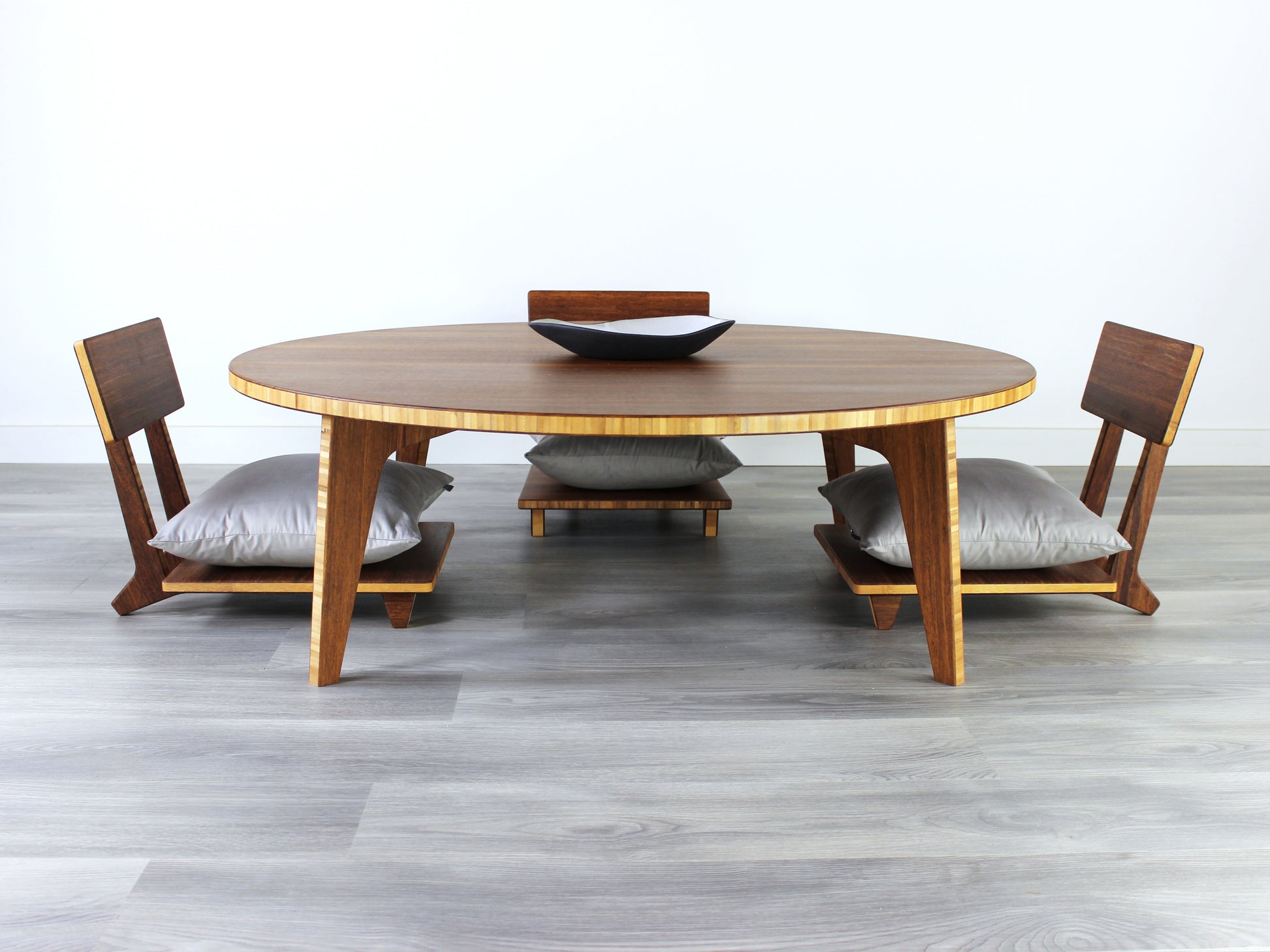 Large Circle Low Table with a Chestnut bamboo finish and midcentury modern design. Three Floor Chair Comfort with grey pillows are set at the table. Small black pottery vessel placed on table top. 

Japandi, minimalist, mid-century, Japanese inspired, modern, contemporary, clean lines, organic design, sustainable bamboo, renewable bamboo, solid bamboo, eco-friendly furniture, natural materials, natural-based finish